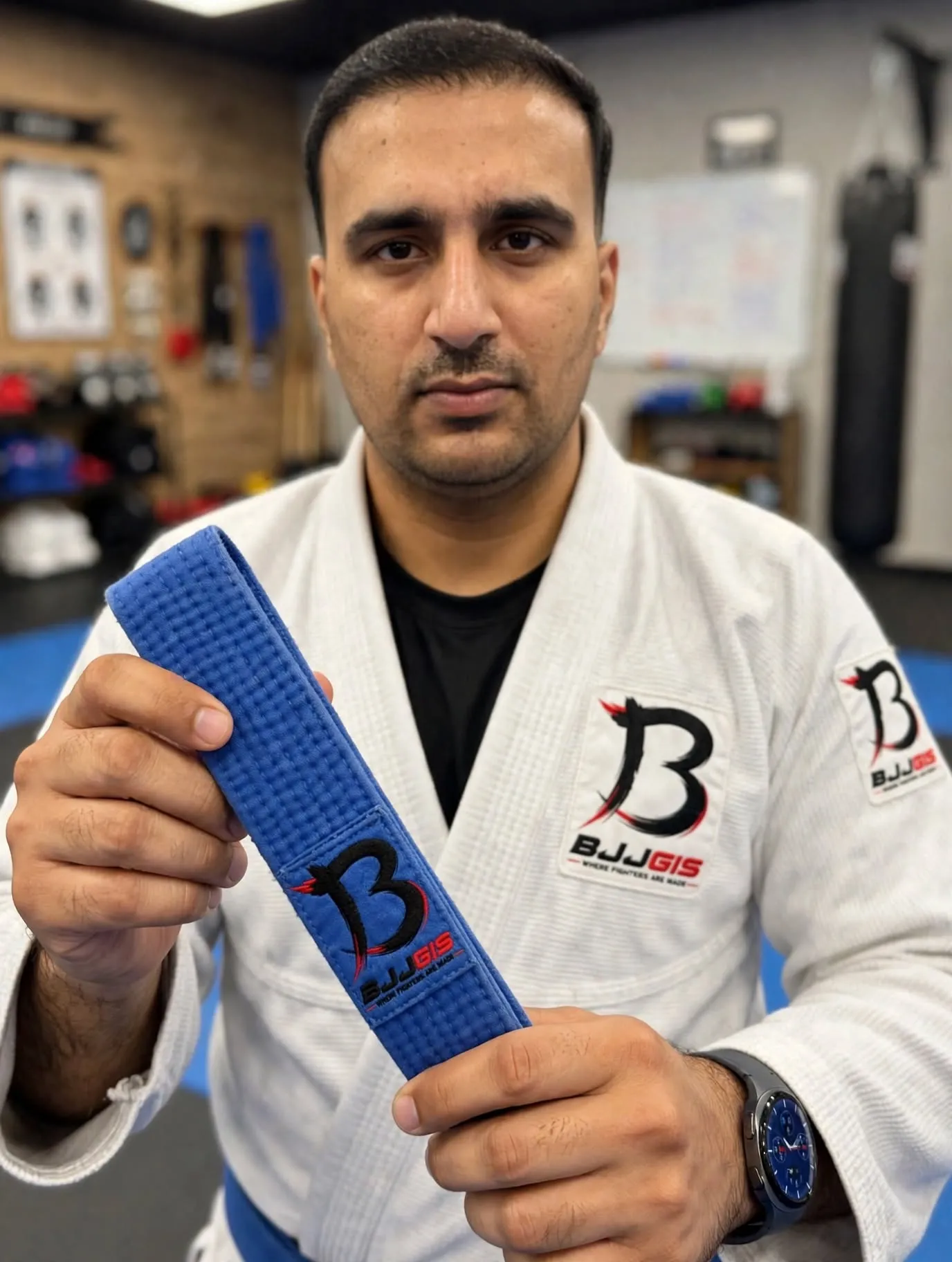 Man wearing white BJJGIS Gi with BJJGIS patches holding blue BJJ belt with large BJJGIS B logo embroidered close-up focus in BJJ gym