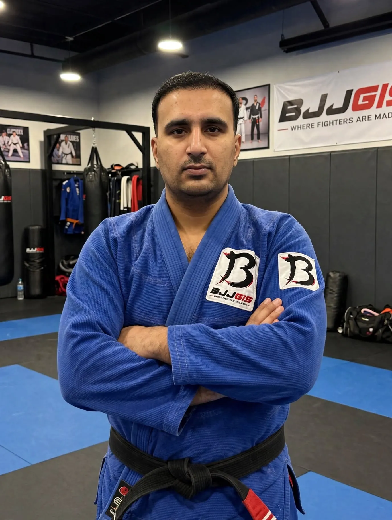 Your model wearing blue BJJGIS BJJ Gi with BJJGIS B logo patches and black belt with red tip arms crossed in BJJ gym with BJJGIS banner