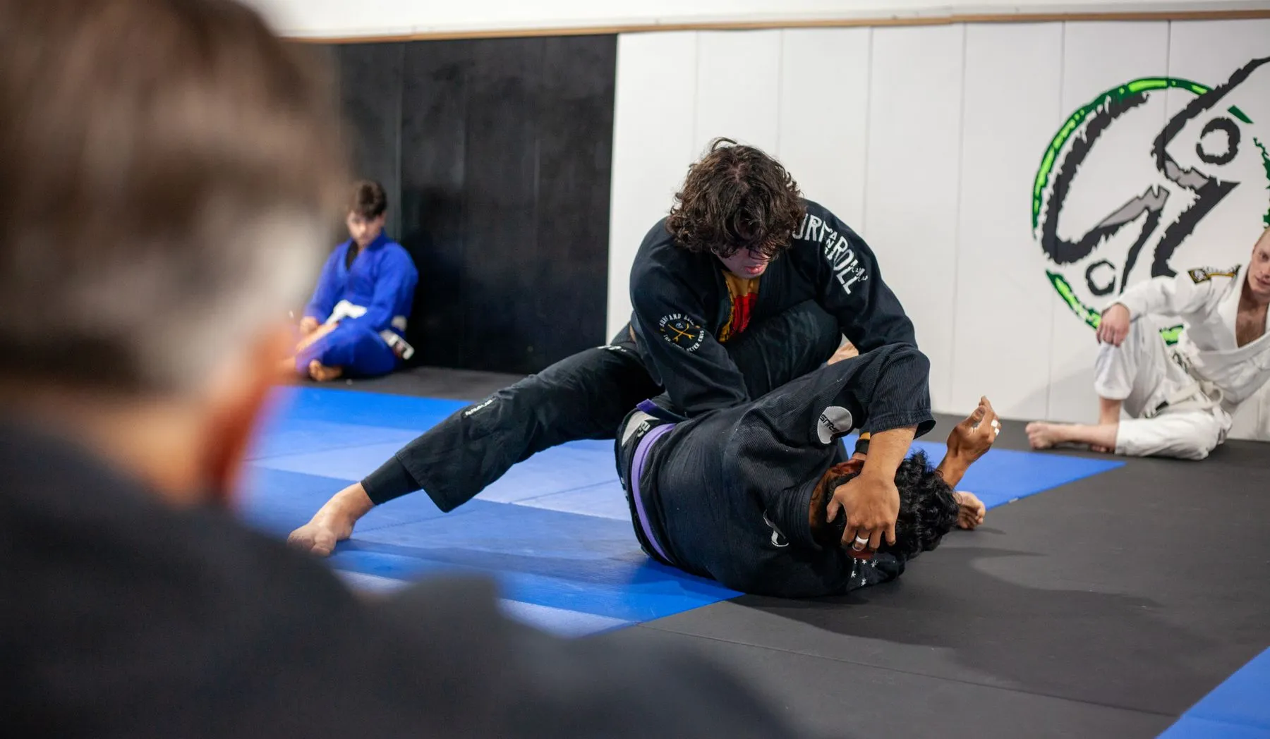 Grappler in a dark gi controlling a training partner on a blue mat